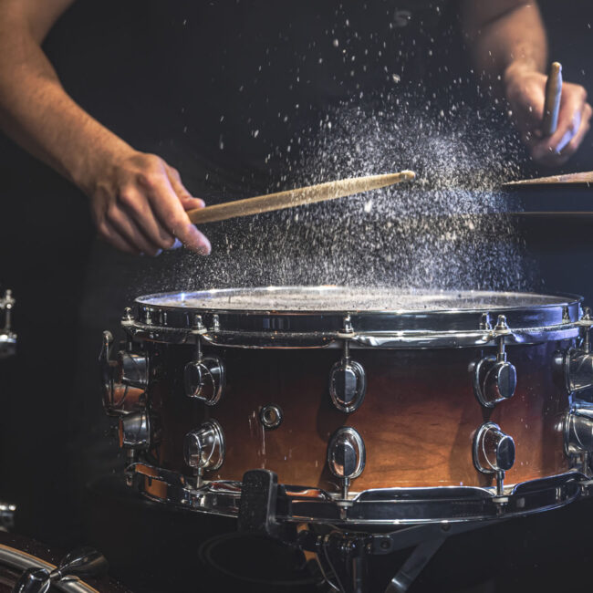 A male drummer plays snare drum with drumsticks in a dark room.